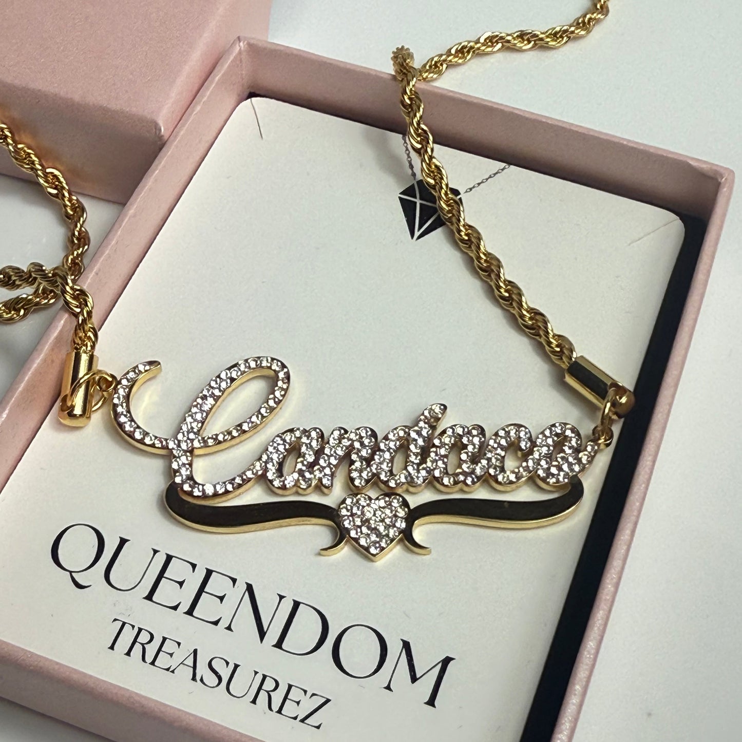 Gold necklace with decorative elements on a 'QUEENDOM TREASUREZ' branded box.