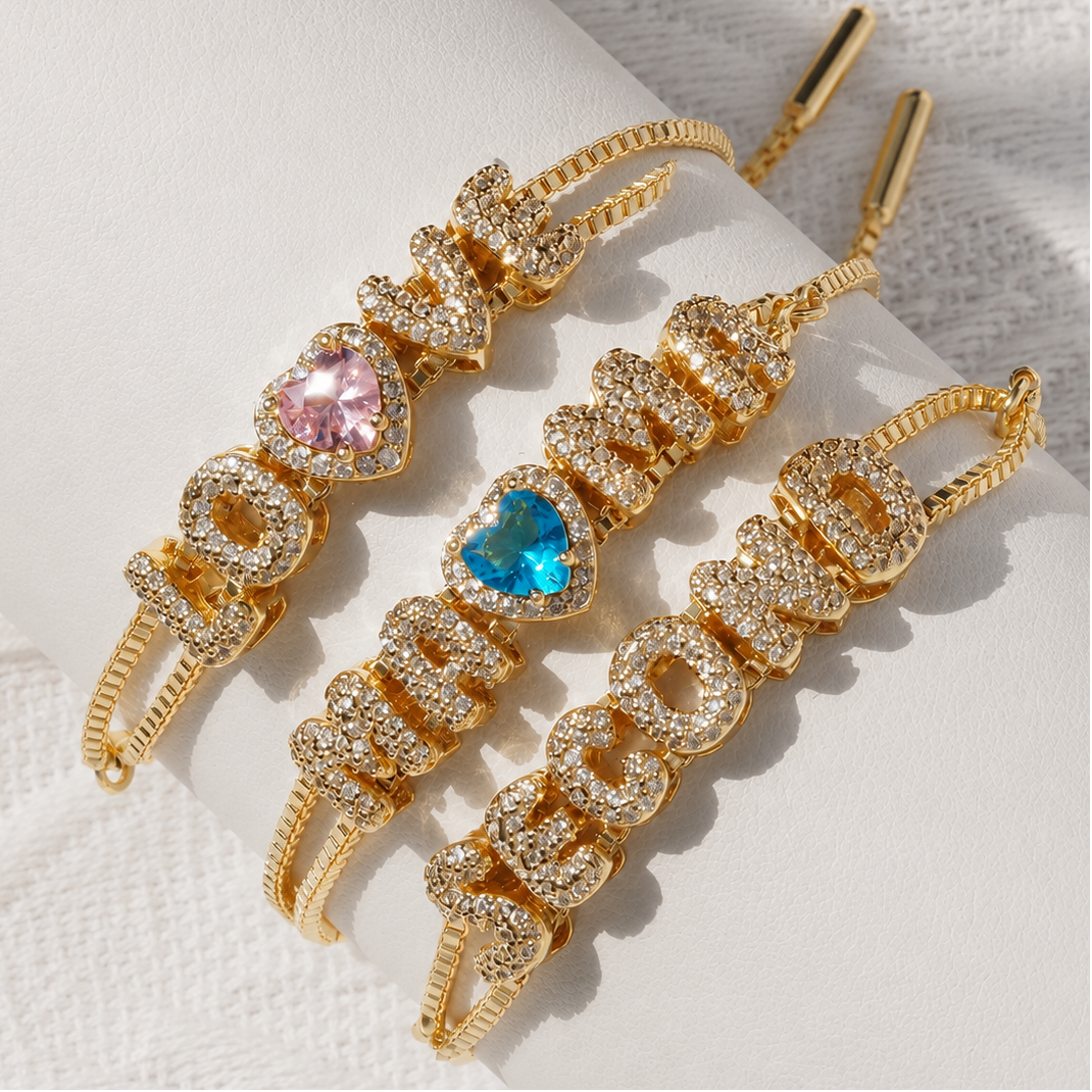 Three gold-plated drop earrings spelling out 'LOVE' vertically, embellished with clear crystals and heart-shaped gemstones in pink and blue, displayed on a white cylindrical stand.