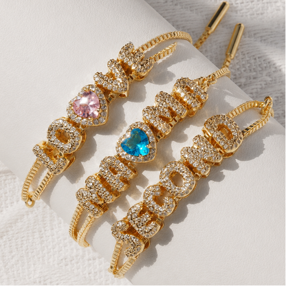 Three gold-plated drop earrings spelling out 'LOVE' vertically, embellished with clear crystals and heart-shaped gemstones in pink and blue, displayed on a white cylindrical stand.