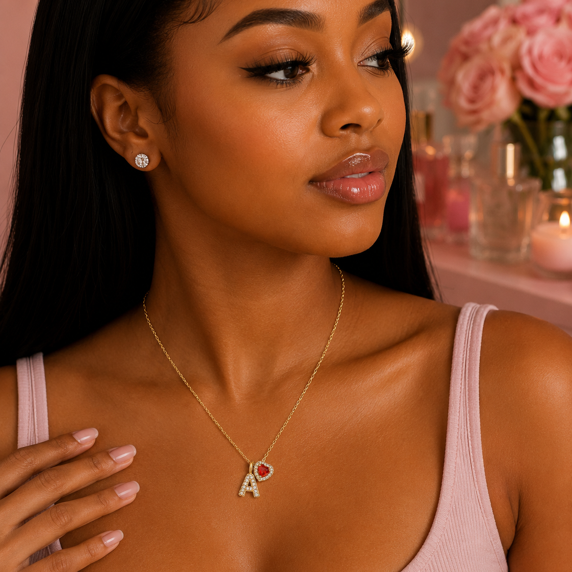 A woman wearing a delicate gold initial necklace with the letter 'A' and a small red heart, paired with a matching stud earring, in a soft pink setting.