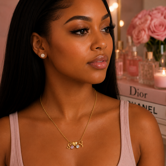A woman wearing a delicate gold necklace with a small cluster of clear and white stones, styled with a matching stud earring. She is wearing a dusty pink ribbed tank top, with a softly blurred background featuring pink roses, perfume bottles, and a book with Dior and Chanel branding.