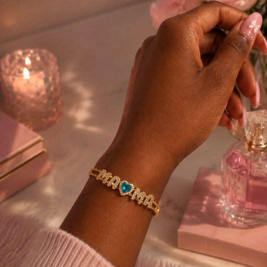 A gold bracelet with a heart-shaped blue gemstone and surrounding small clear stones, worn on a person's wrist in a pink-themed setting with roses and a perfume bottle.