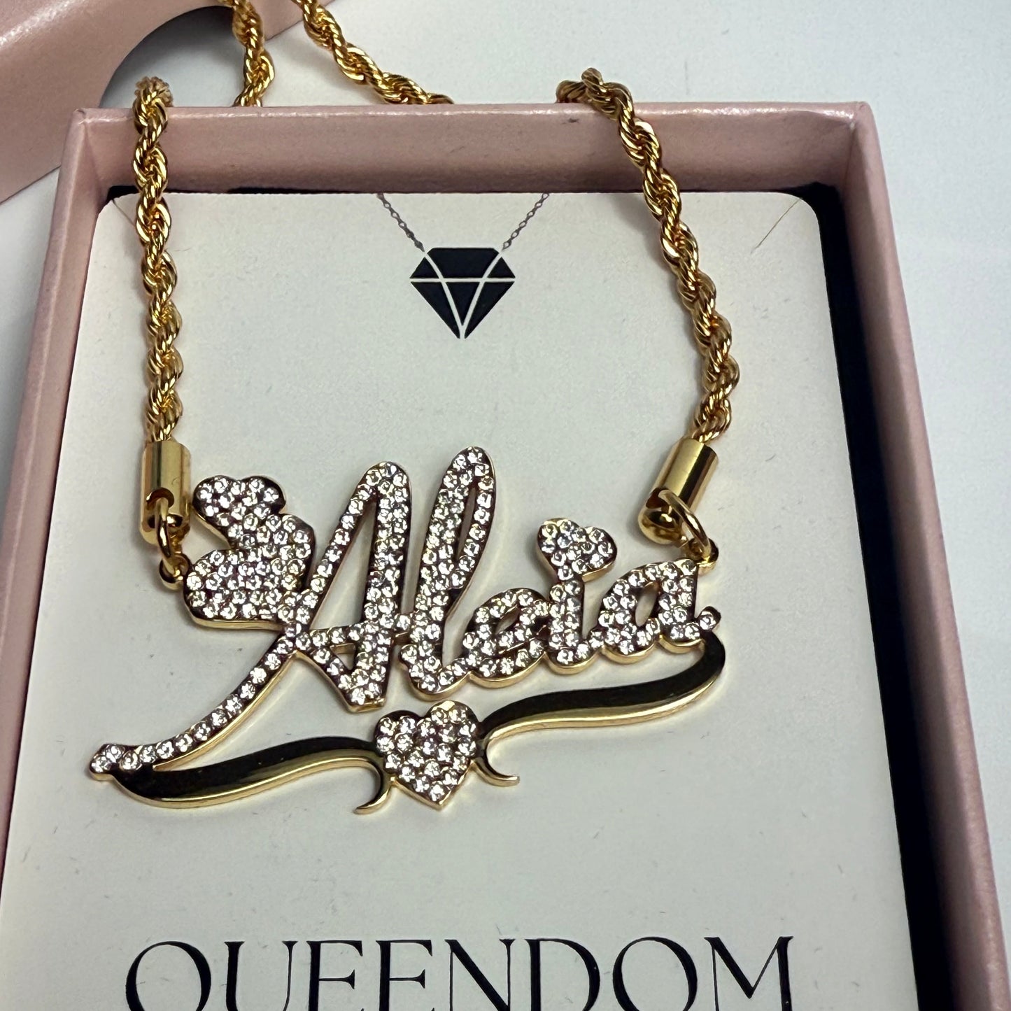 Gold necklace with intricate design on a card labeled 'QUEENDOM TREASUREZ' in a pink box.