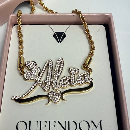 Gold necklace with intricate design on a card labeled 'QUEENDOM TREASUREZ' in a pink box.