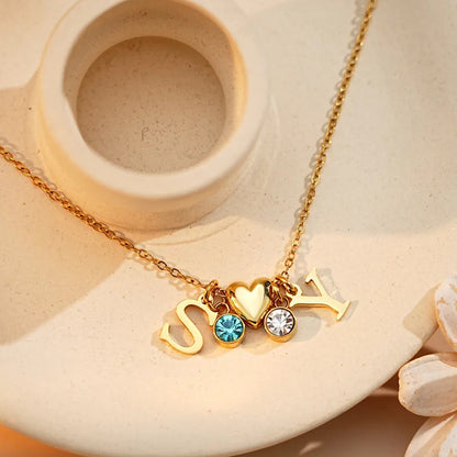 Gold initial necklace with three charms (letter S, heart, letter Y) and two round crystal rhinestones, displayed on a beige ceramic dish with a flower prop.