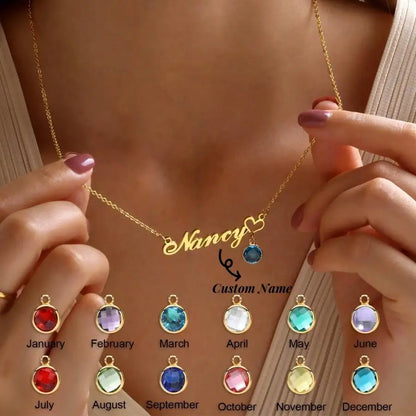 gold necklace with a script nameplate reading 'Nancy' and a small heart-shaped charm. The image includes a row of color options for the nameplate and charm stones at the bottom, labeled by months.