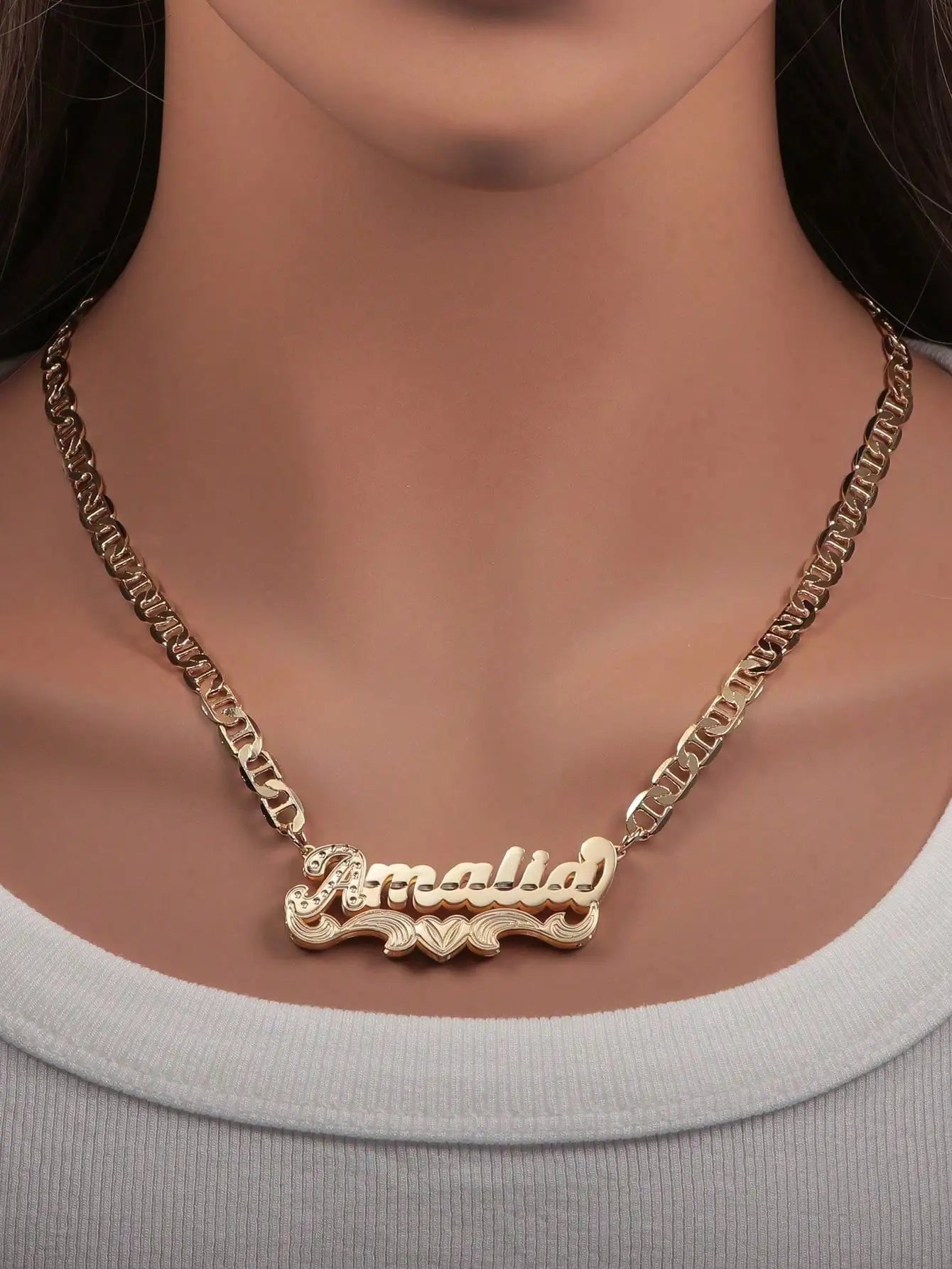 A gold-tone name necklace with the name 'Amalid' in cursive script and small heart details, shown worn on a model's neck over a white ribbed top.