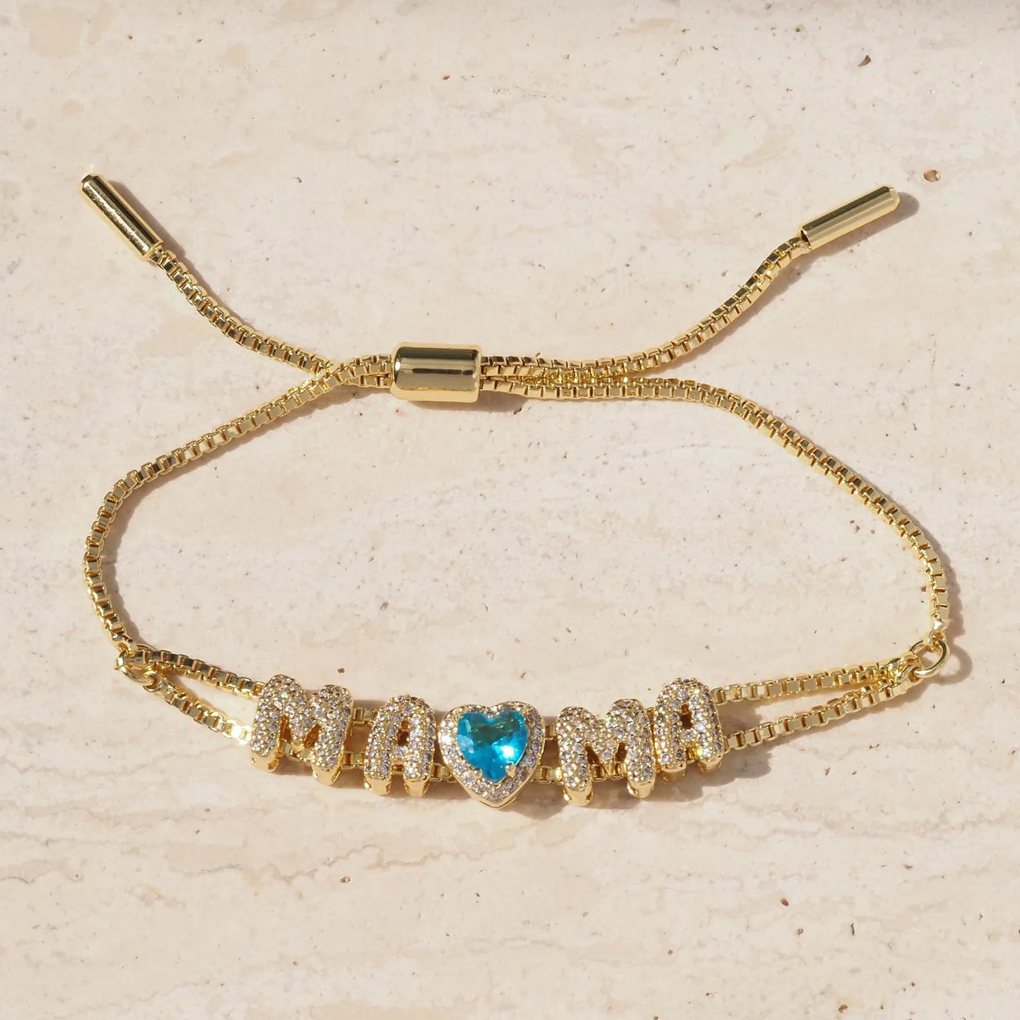 A gold-tone adjustable cord bracelet featuring the word 'MAMA' spelled out in sparkling pave-set letters, with a central heart-shaped blue gemstone, displayed on a light beige textured surface.