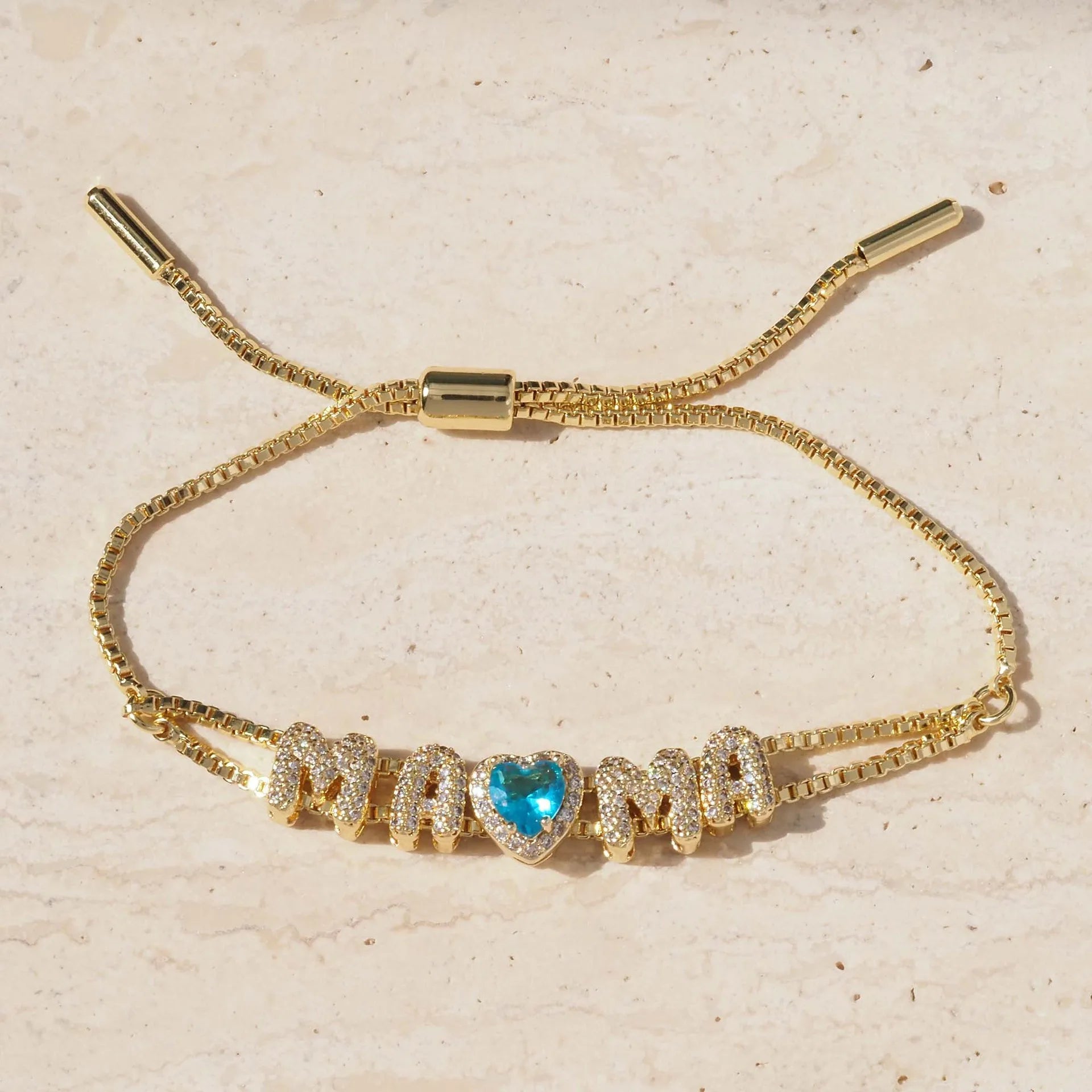 A gold-tone adjustable cord bracelet featuring the word 'MAMA' spelled out in sparkling pave-set letters, with a central heart-shaped blue gemstone, displayed on a light beige textured surface.